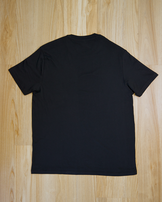 The Ecotton Everyday Tee Black Three pack lying flat on a light wood floor, viewed from the back. The smooth fabric and simple design convey a minimalistic style.